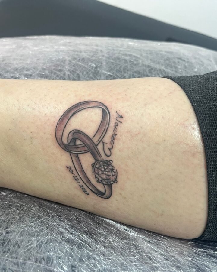 Tatuaje realista de anillos entrelazados con nombre y fecha en la pierna