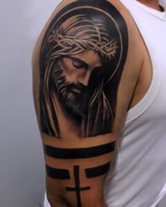 Tatuaje de Cristo con corona de espinas en realismo religioso en el hombro, con bandas en negro y cruz