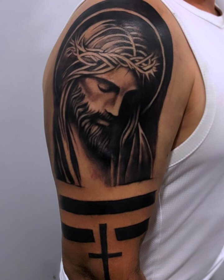 Tatuaje de Cristo con corona de espinas en realismo religioso en el hombro, con bandas en negro y cruz