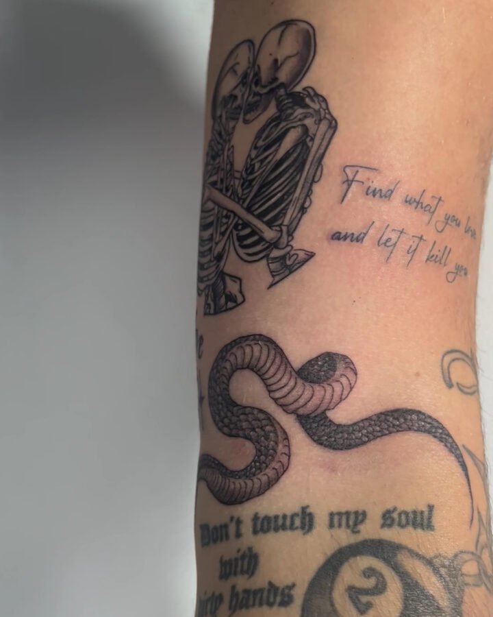 Tatuajes en negro de esqueletos abrazados, serpiente y frase en el brazo