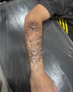 Tatuaje fine line de flores de loto con nombre y año en lettering en el antebrazo